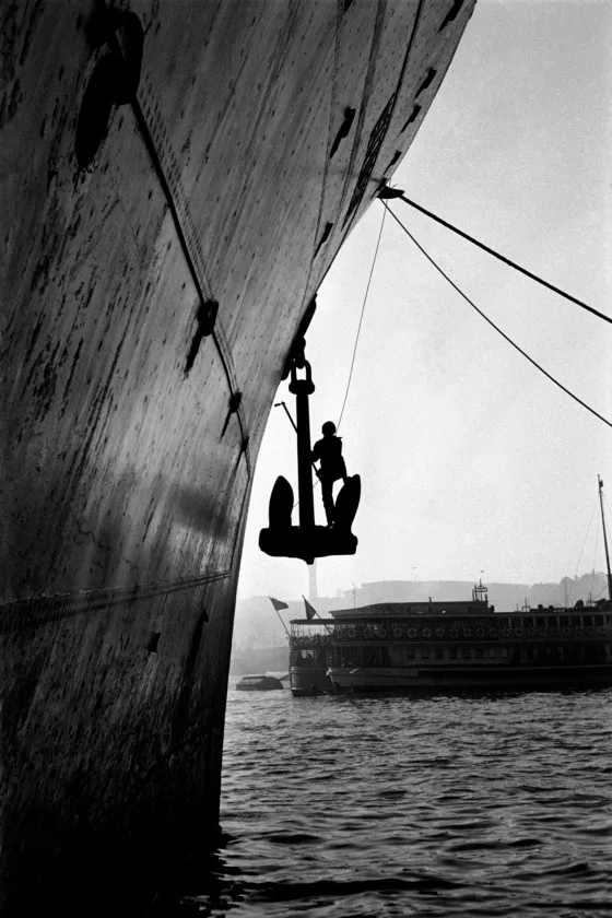 The bow and anchor of a ship, with a boat heading for the horizon behind it.